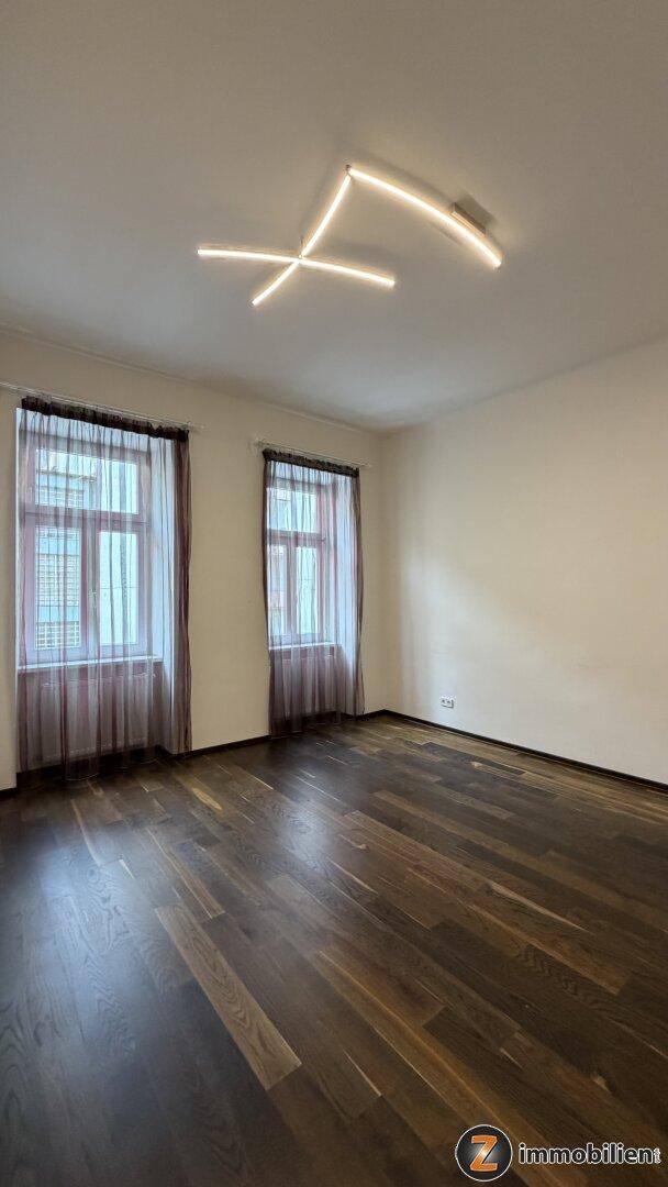 Heller Raum mit modernem Deckenlicht, dunklem Holzboden und zwei Fenstern.