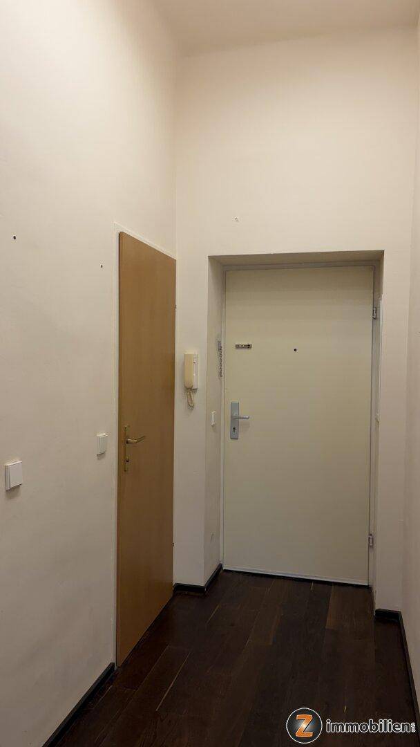 Heller Flur mit dunklem Holzboden und zwei Türen, darunter die Eingangstür der Wohnung.