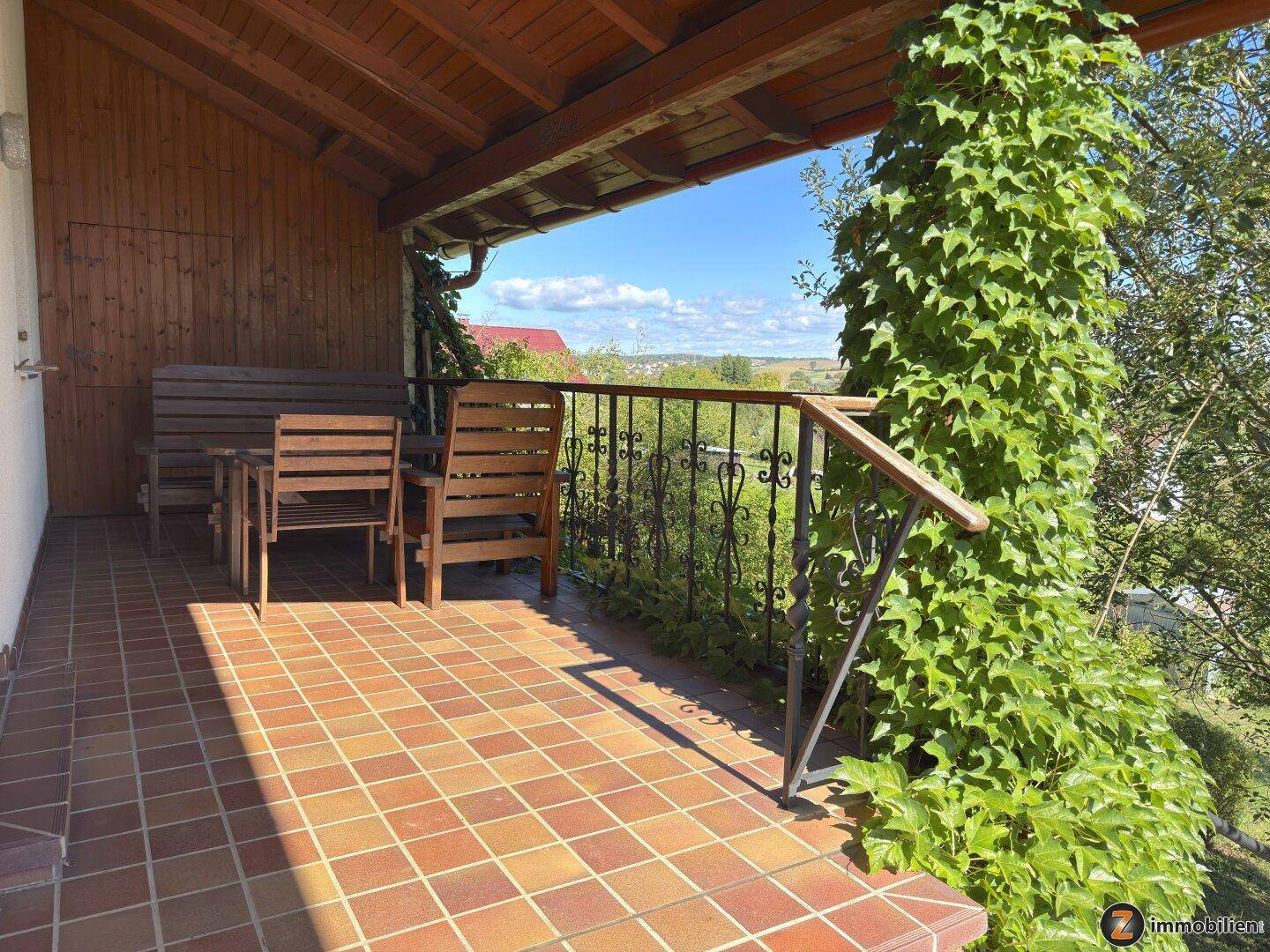 Überdachter Balkon mit Holzmöbeln, Fliesenboden und weitem Blick in die Landschaft.
