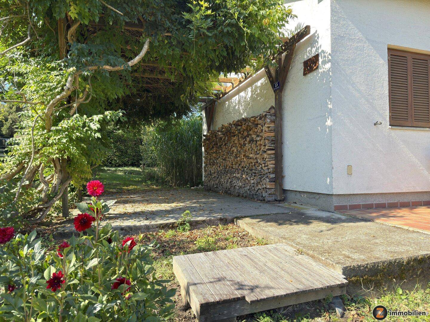Seitenansicht des Hauses mit gestapeltem Brennholz und blühenden Blumen im Vordergrund.