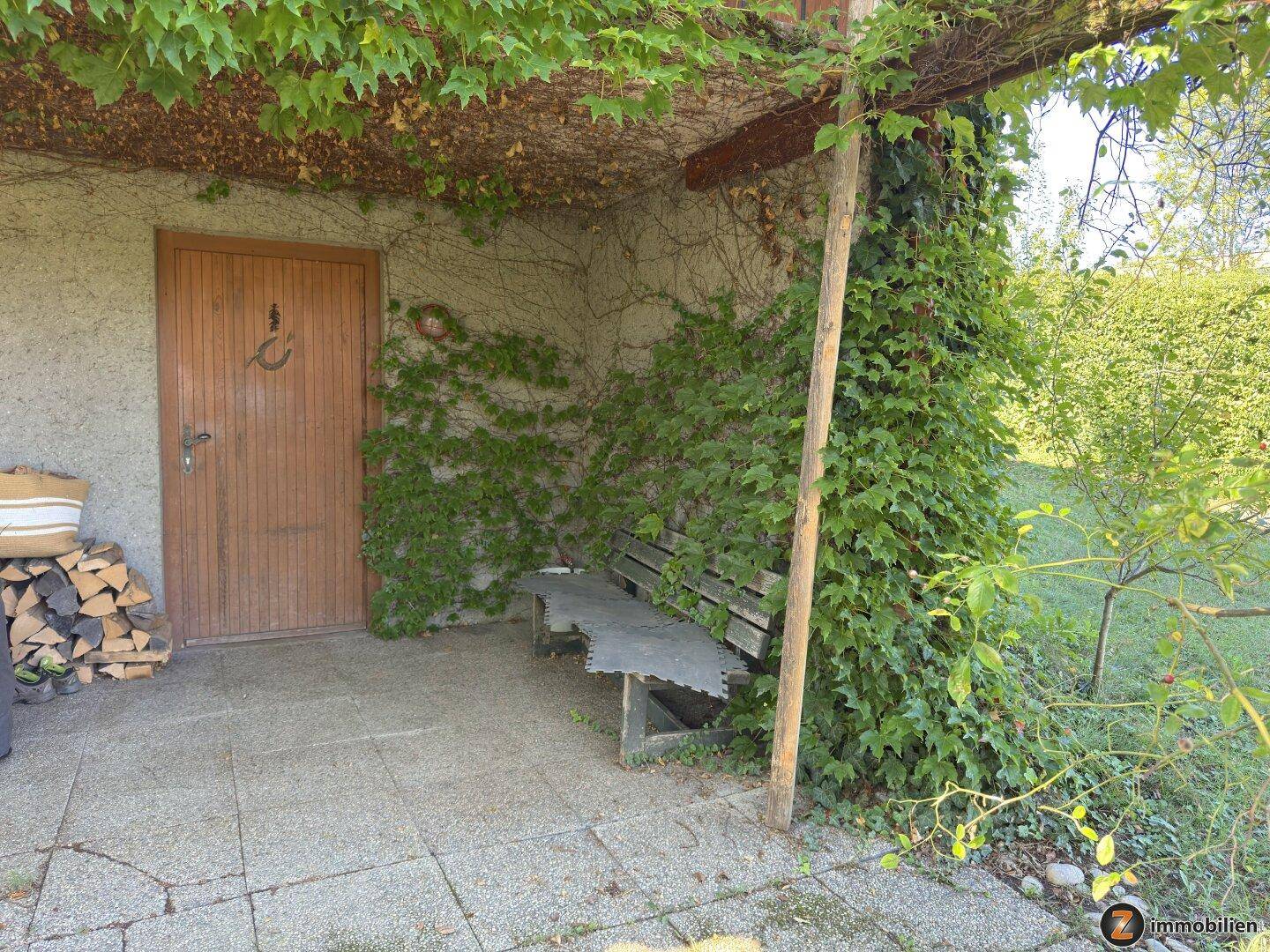 Überdachter Eingangsbereich mit Sitzbank und üppigem Grün an der Hauswand.