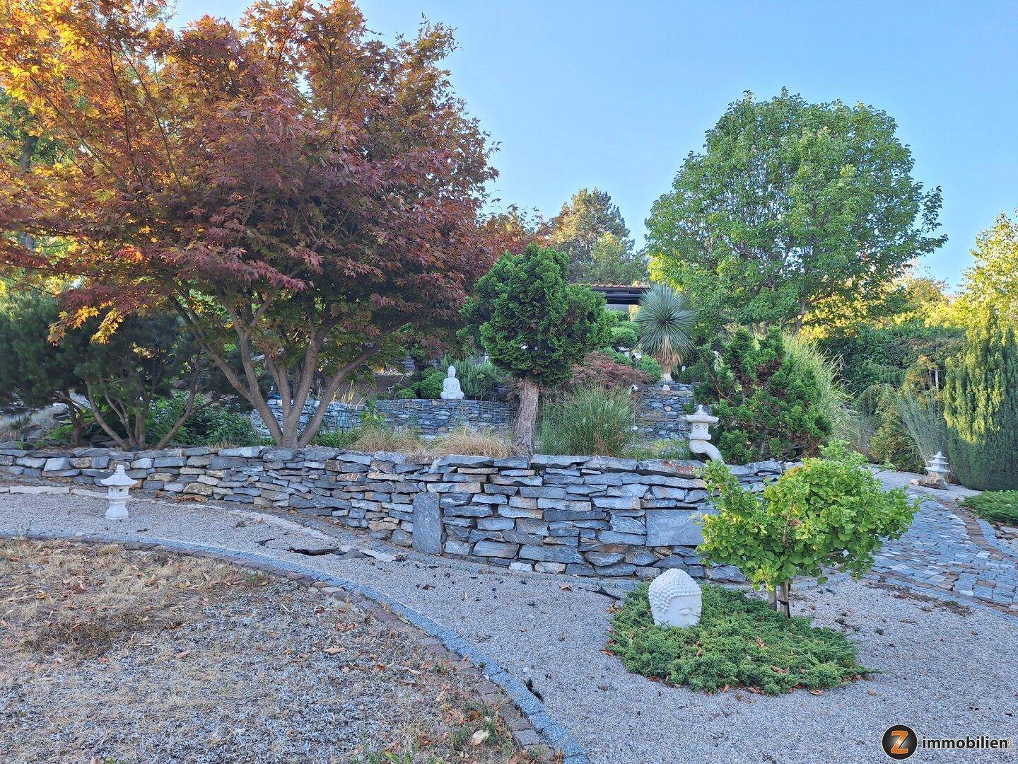 Ruhiger Zen-Garten mit Kieswegen, Steinmauern, verschiedenen Bäumen und dekorativen Buddha-Statuen.