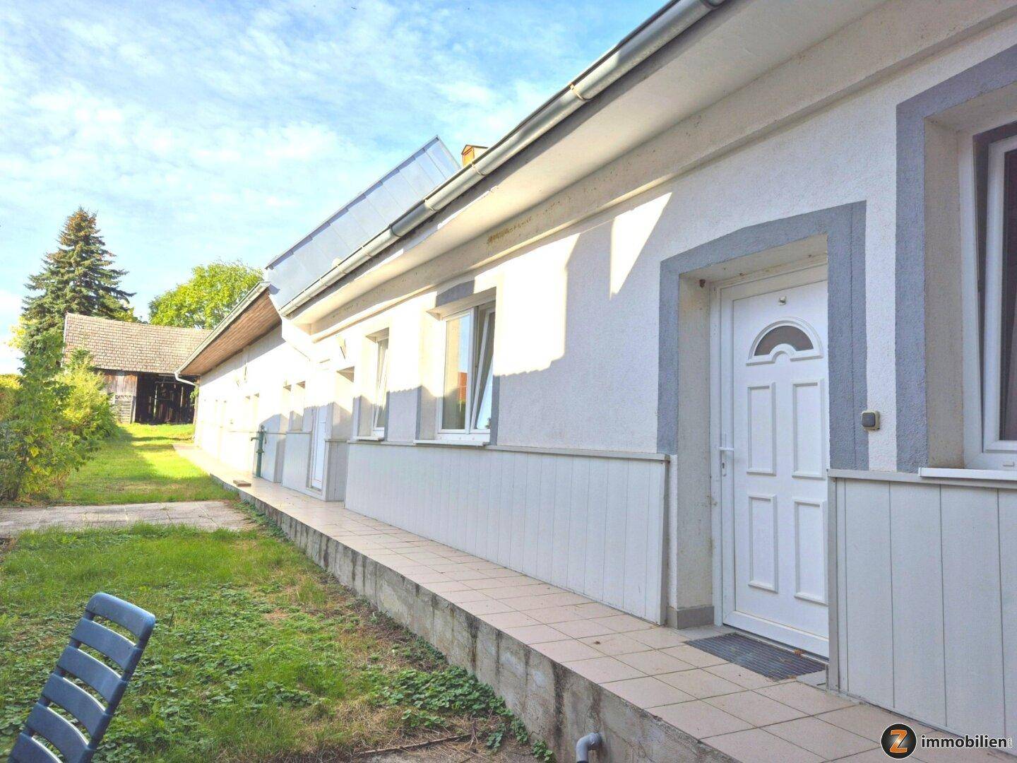Seitenansicht des Hauses mit Eingangstür, Fenstern und einem gepflasterten Weg entlang der Fassade.
