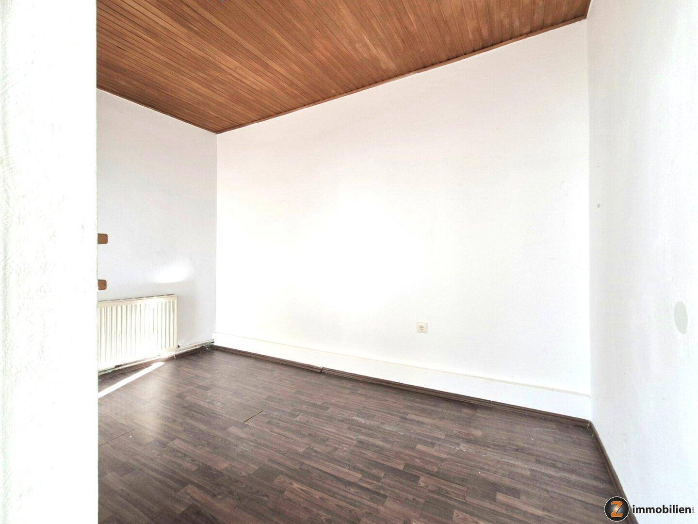 Leeres Zimmer mit dunklem Laminatboden, Holzdeckenverkleidung und Heizkörper unter dem Fenster.