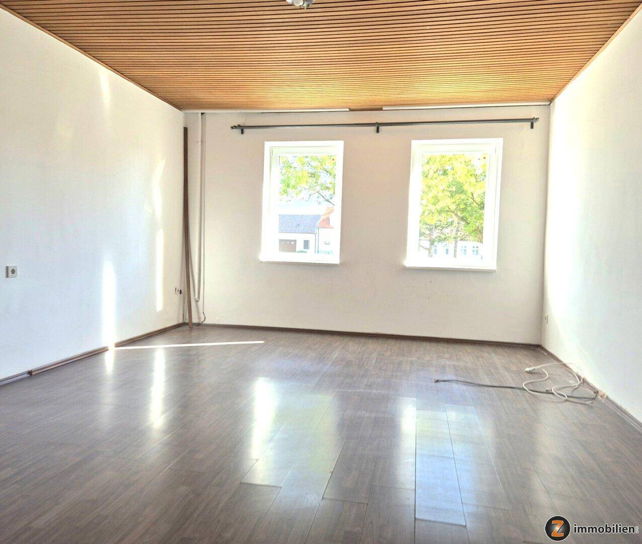 Helles, leeres Zimmer mit Holzdeckenverkleidung, Laminatboden und zwei Fenstern mit Blick ins Grüne.