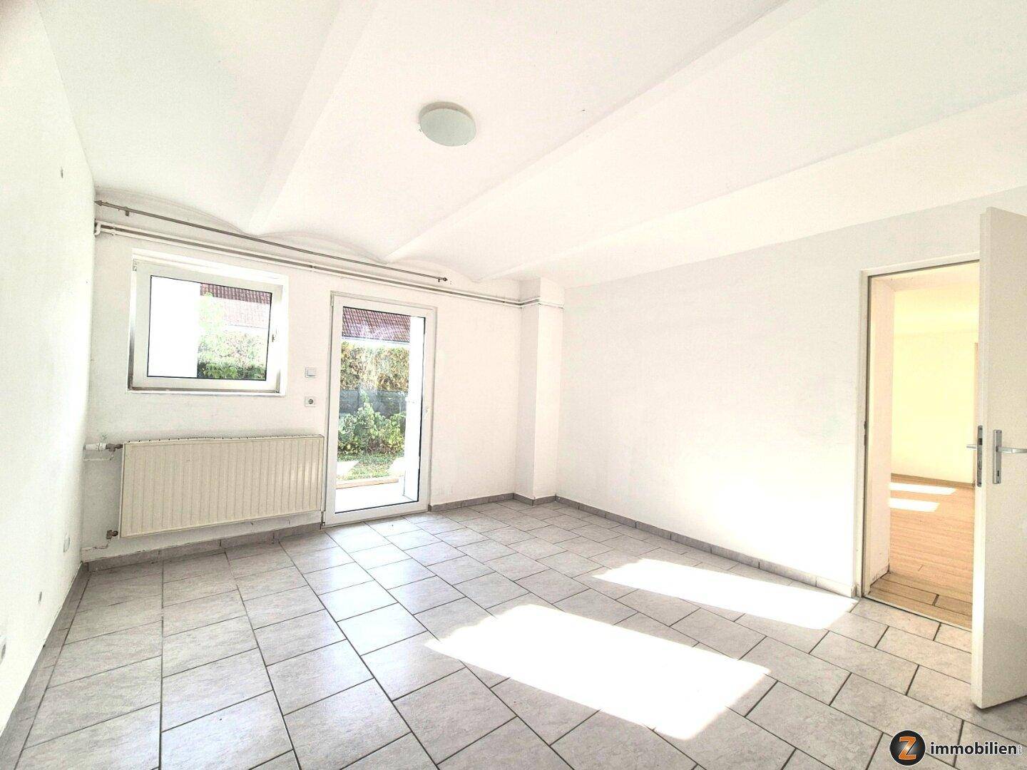Helles Zimmer mit Fliesenboden und direktem Zugang zum Garten durch eine große Glastür.