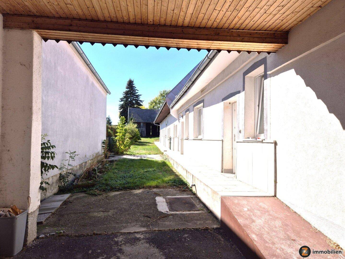Überdachter Außenbereich entlang der Gebäudeseite mit Blick auf den Garten und das Nachbargebäude.