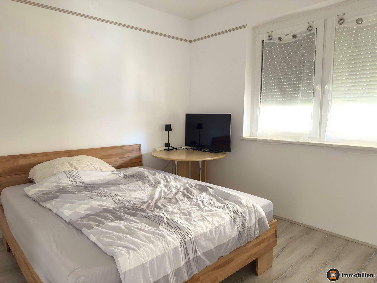 Helles Schlafzimmer mit Einzelbett, Fernseher und Fenster mit Rollladen.