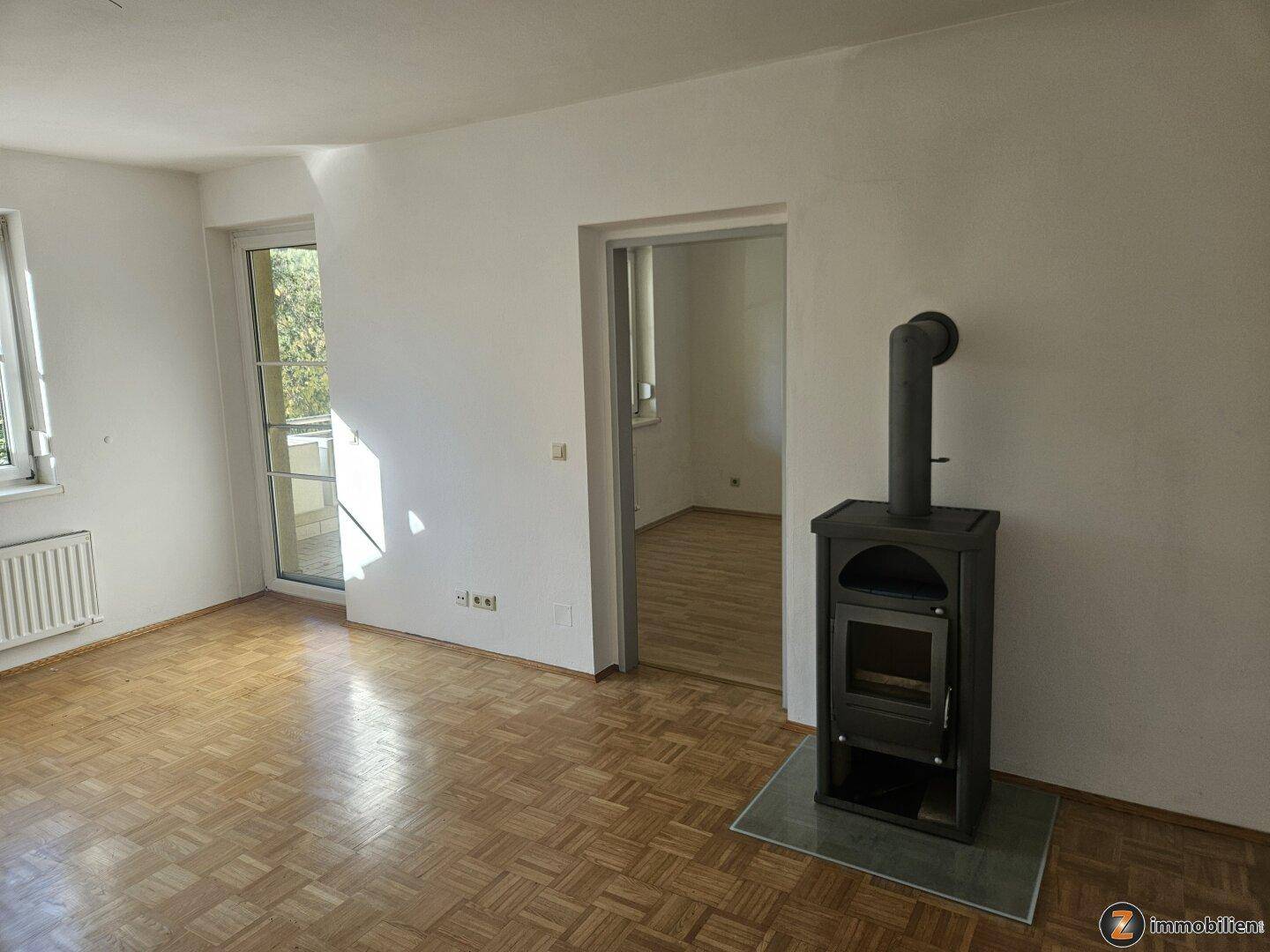 Wohnzimmer mit Parkettboden, Holzofen und Zugang zu einem sonnigen Außenbereich.