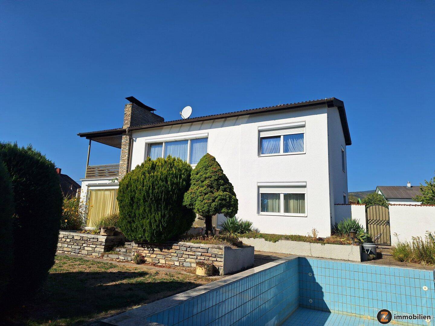 Einfamilienhaus mit weißer Fassade, Rollläden und einem leeren Swimmingpool im Garten.