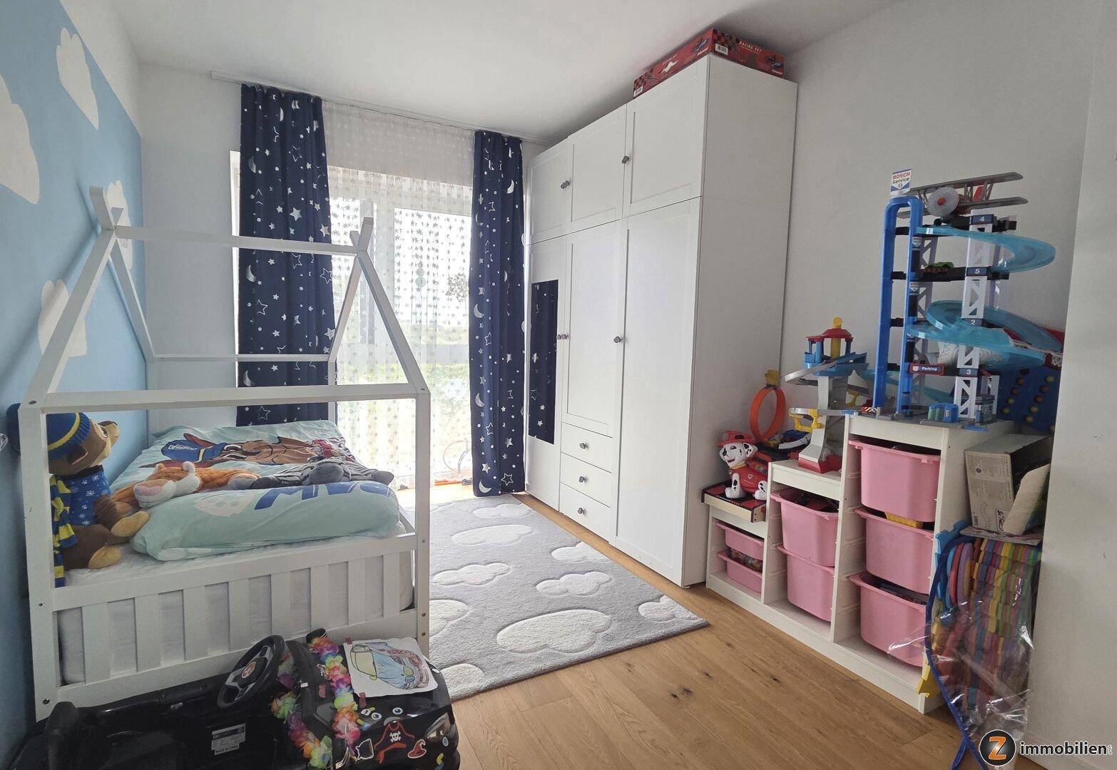 Kinderzimmer mit einem Hausbett, weißem Kleiderschrank und Spielzeug, dekoriert mit blauen Wolken.