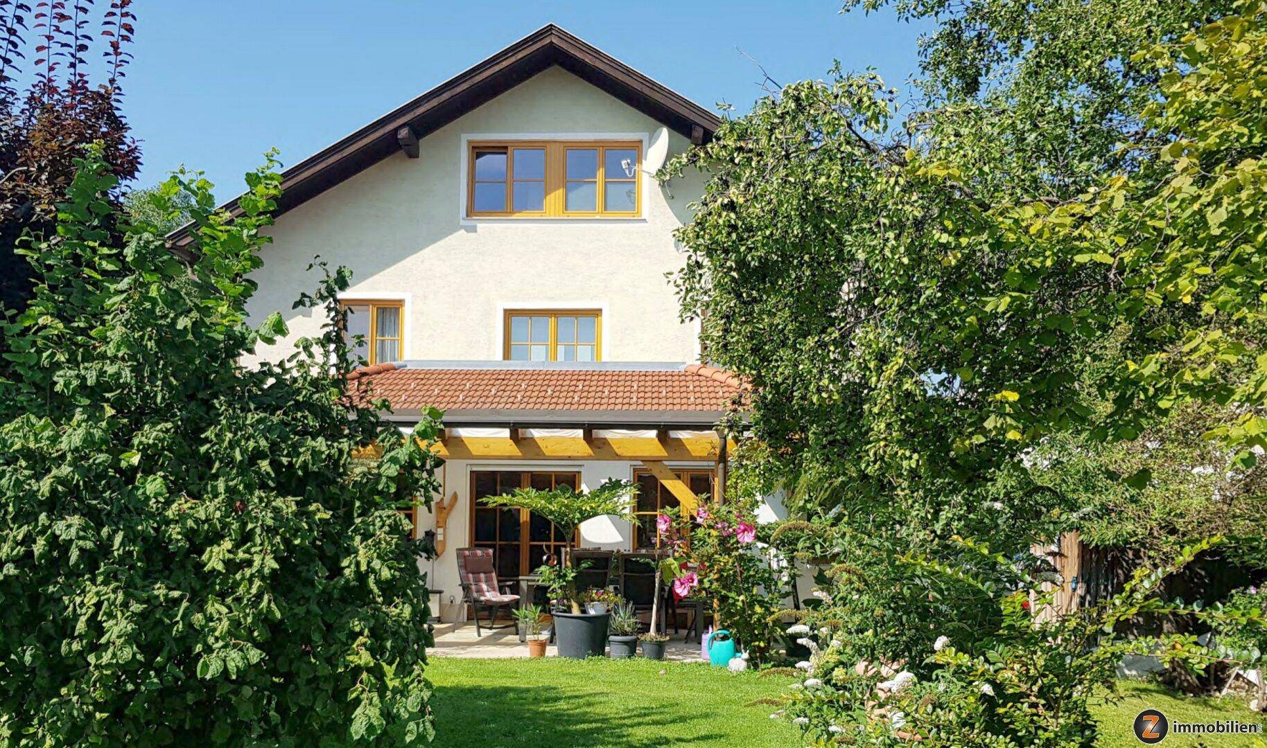 Hausfassade mit Garten, Terrasse und üppiger Bepflanzung unter blauem Himmel.