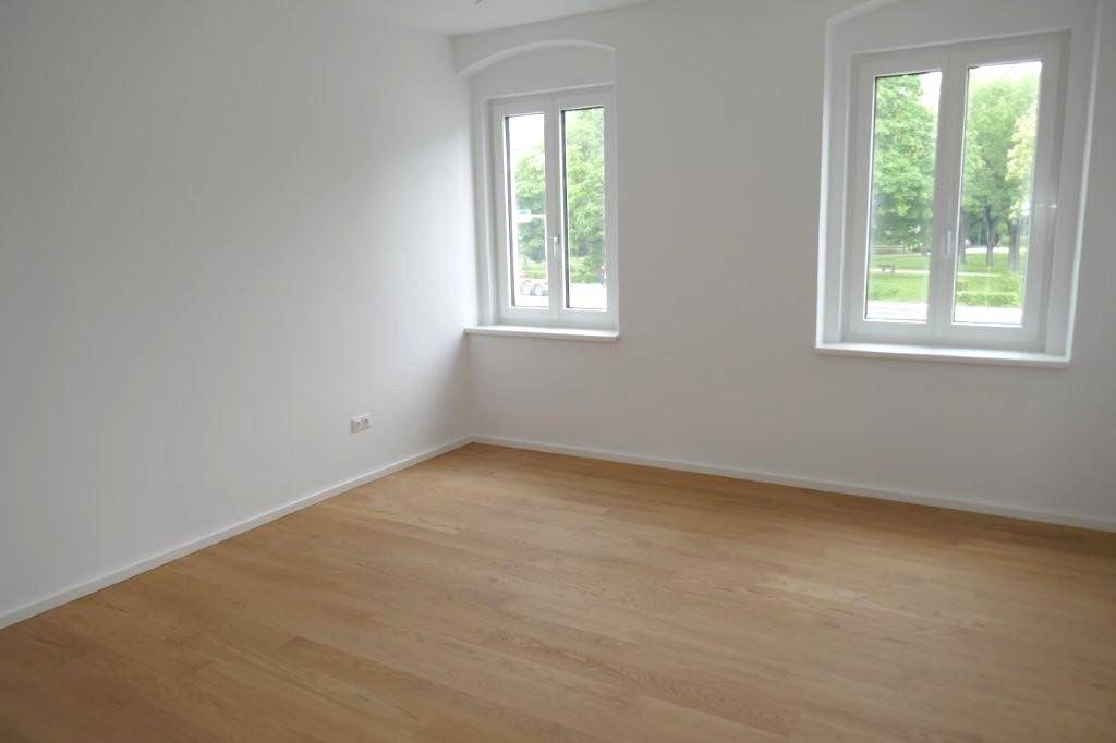 Leeres Zimmer mit zwei Fenstern, hellem Holzfußboden und weiß gestrichenen Wänden.