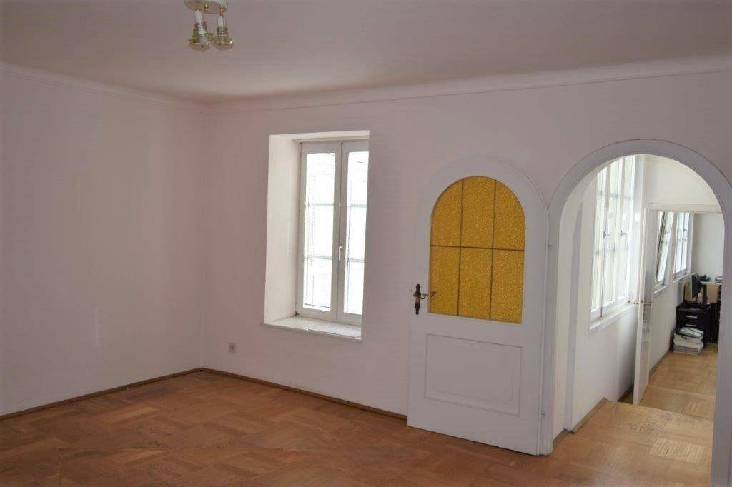 Heller Raum mit Parkettboden, einem Fenster und einer Tür mit gelbem Glaseinsatz, verbunden durch einen Bogen.