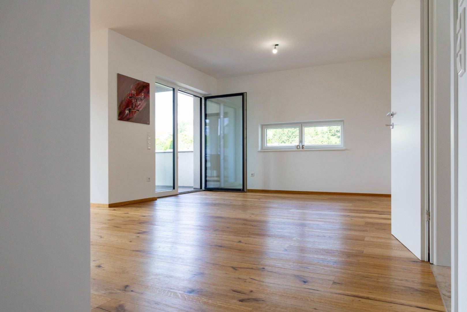 Geräumiger Wohnbereich mit warmem Holzboden und viel Tageslicht durch große Fenster.