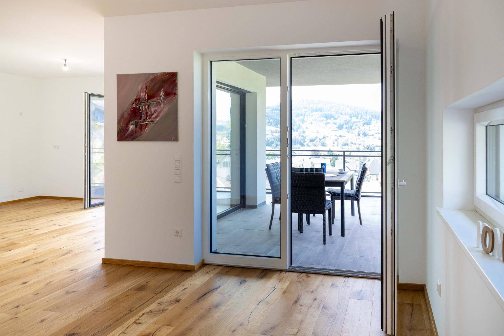 Geräumiges Wohnzimmer mit Holzboden und großen Glasfronten, die zu einem möblierten Balkon führen.