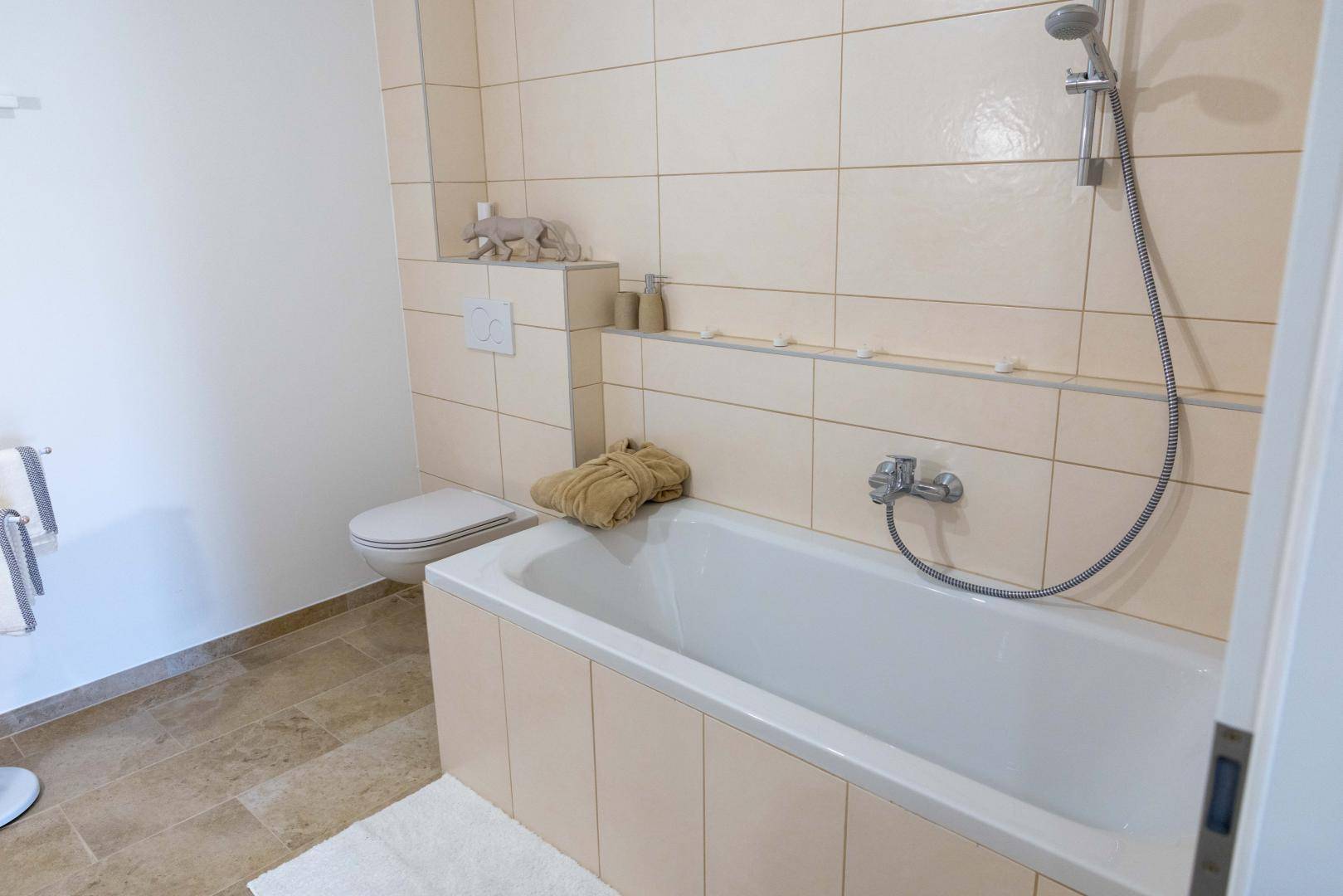 Helles Badezimmer mit beigefarbenen Fliesen, Badewanne und wandmontiertem WC.