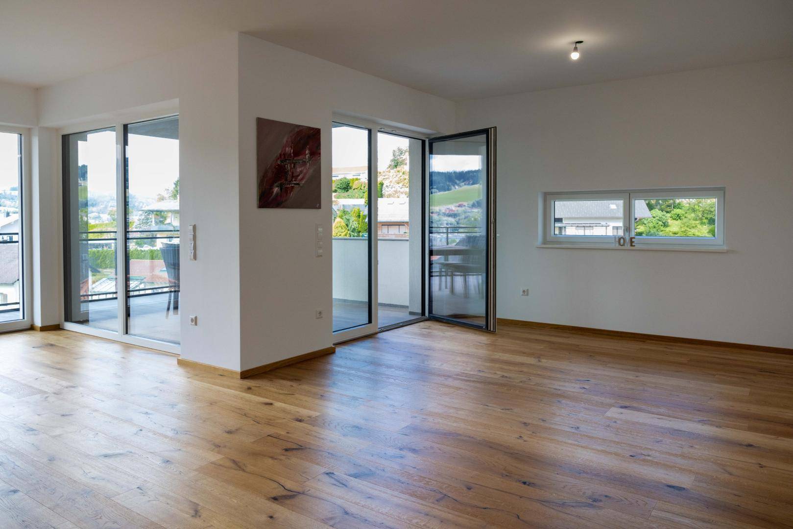 Geräumiger Wohnbereich mit Holzboden, großen Fenstern und Zugang zu einem Balkon.