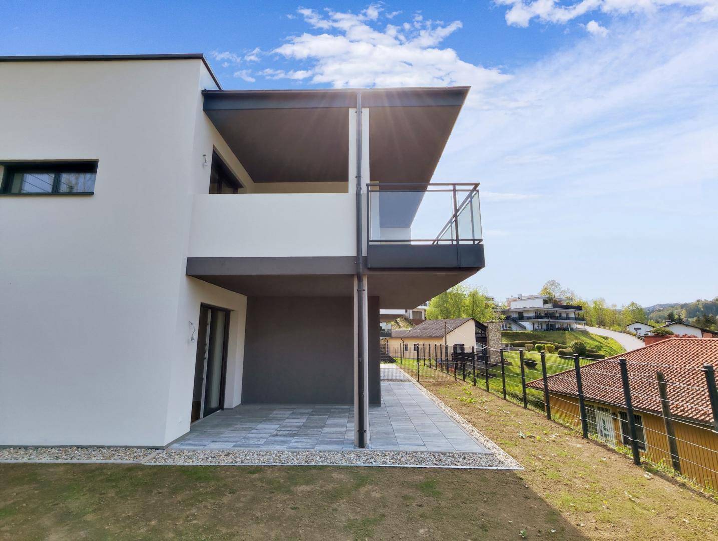 Seitliche Ansicht des Neubaus mit einer gepflasterten Terrasse und Balkon im Obergeschoss.