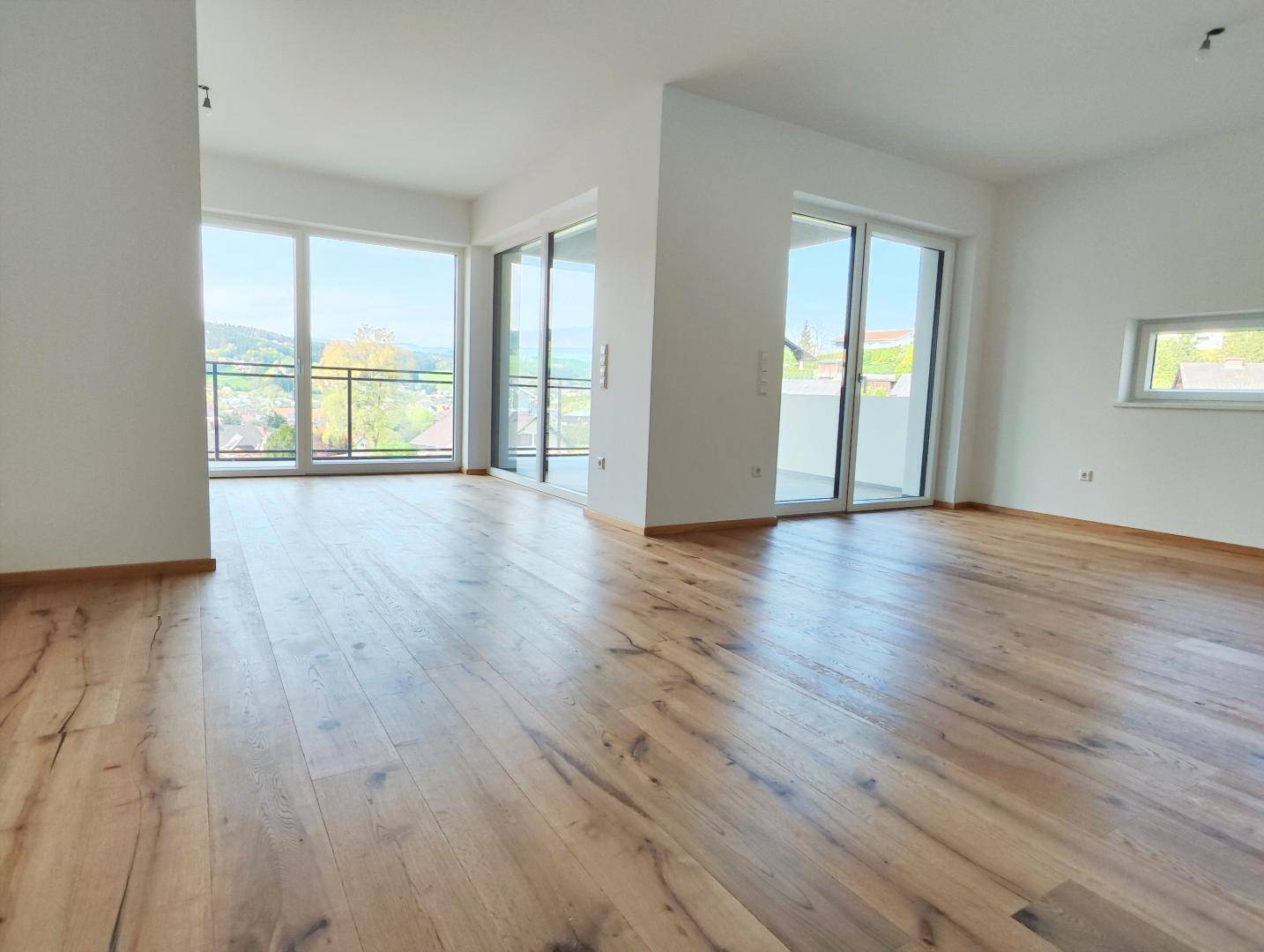 Lichtdurchfluteter Wohnraum mit großen Fenstern, Holzboden und Zugang zu einem Balkon.
