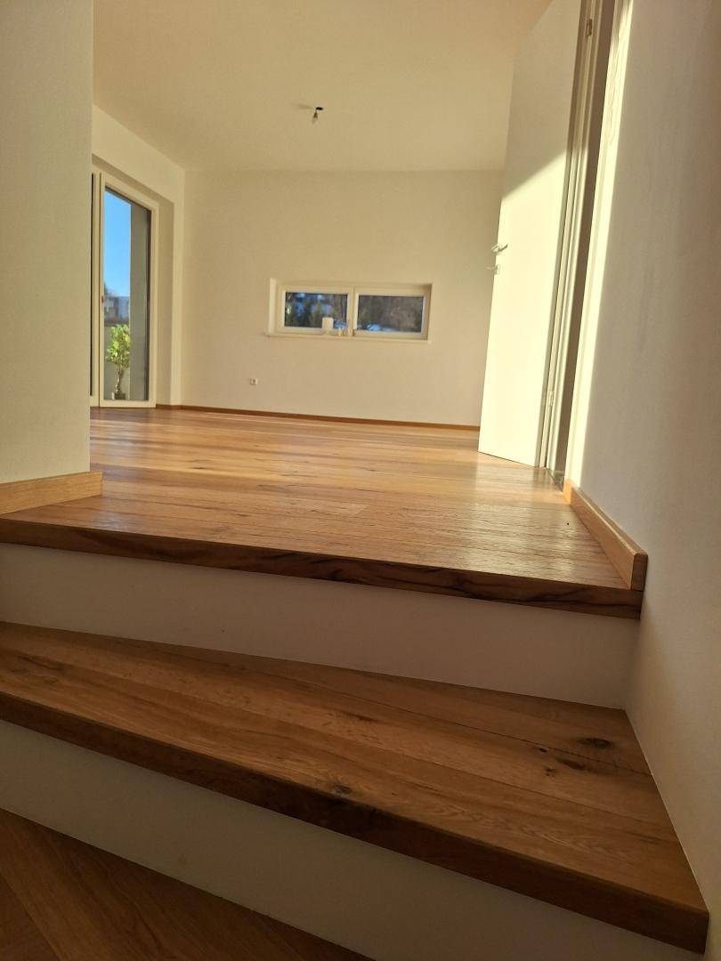 Holztreppe führt zu einem hellen Raum mit Holzboden und Fenstern, sonnendurchflutet.