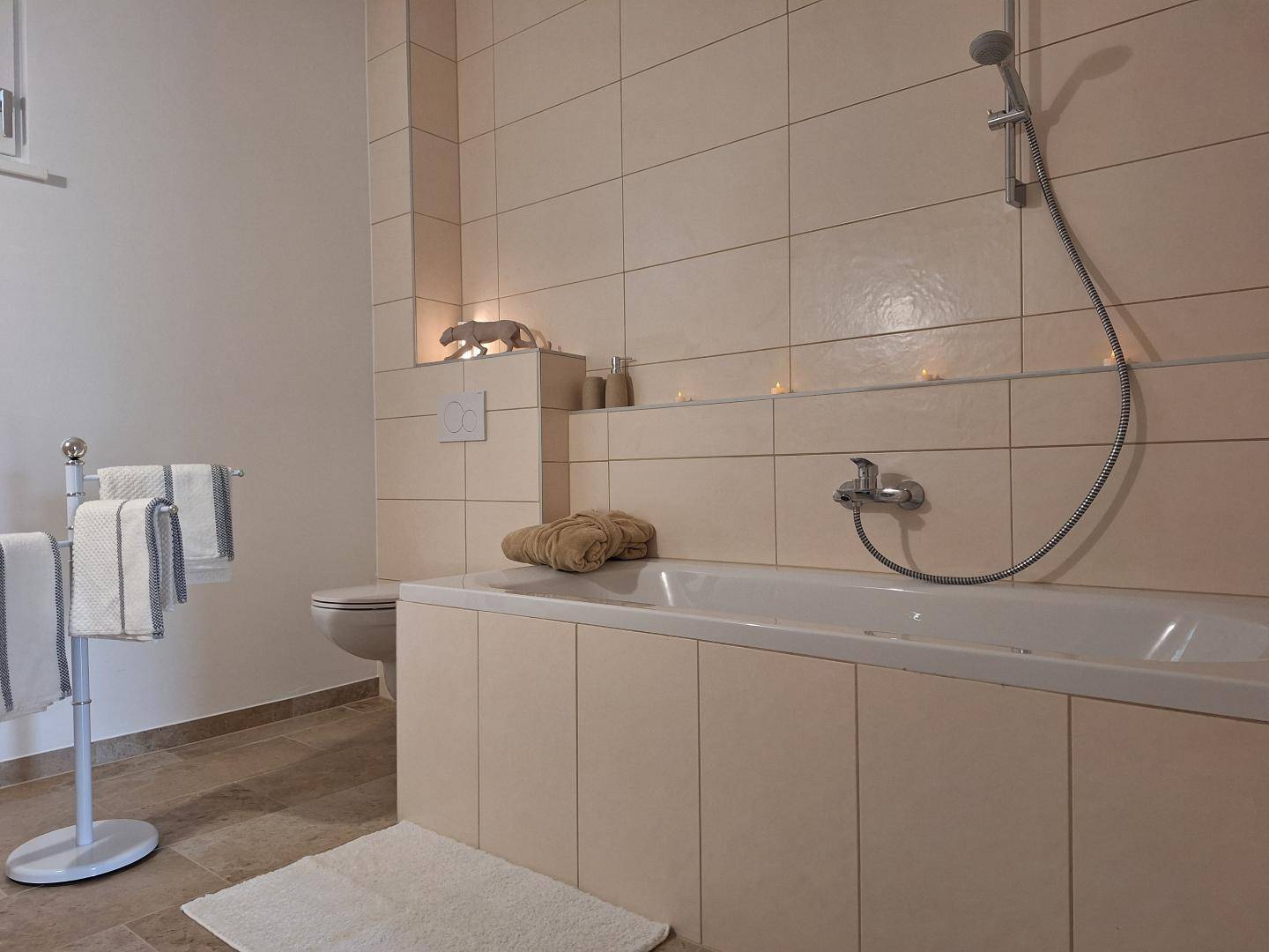 Helles Badezimmer mit Badewanne, beigefarbenen Fliesen und einem Handtuchhalter.