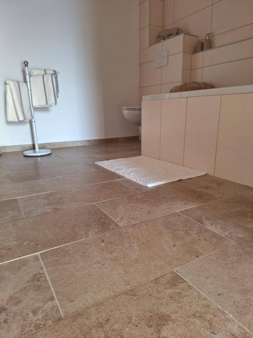 Badezimmerboden mit großformatigen beigefarbenen Fliesen und einem Badteppich.