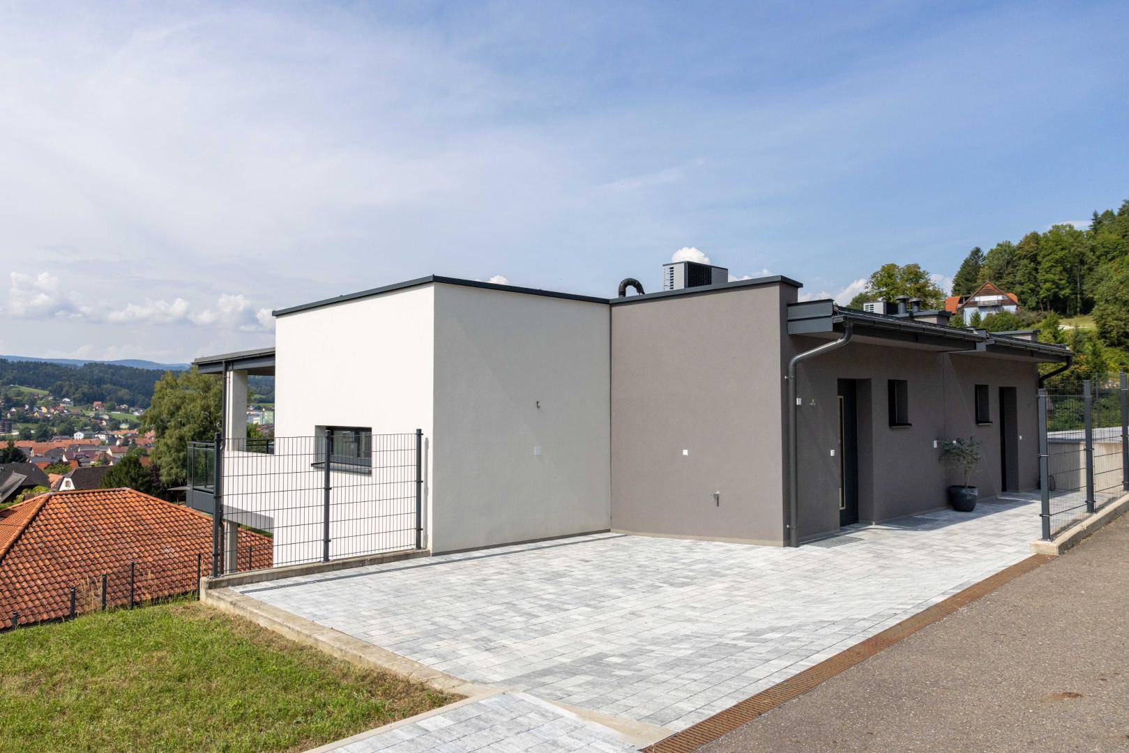 Moderne Gebäudefassade mit gepflastertem Vorplatz und Parkmöglichkeiten in Hanglage.