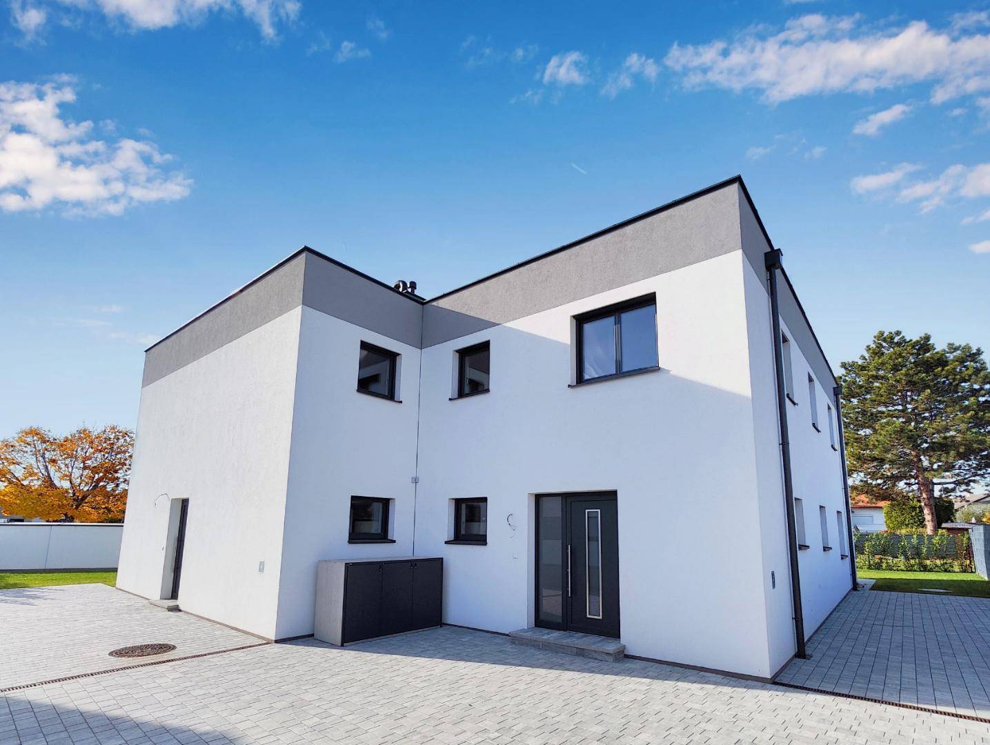 Moderne Hausfassade mit klaren Linien und gepflastertem Eingangsbereich unter strahlendem Himmel.