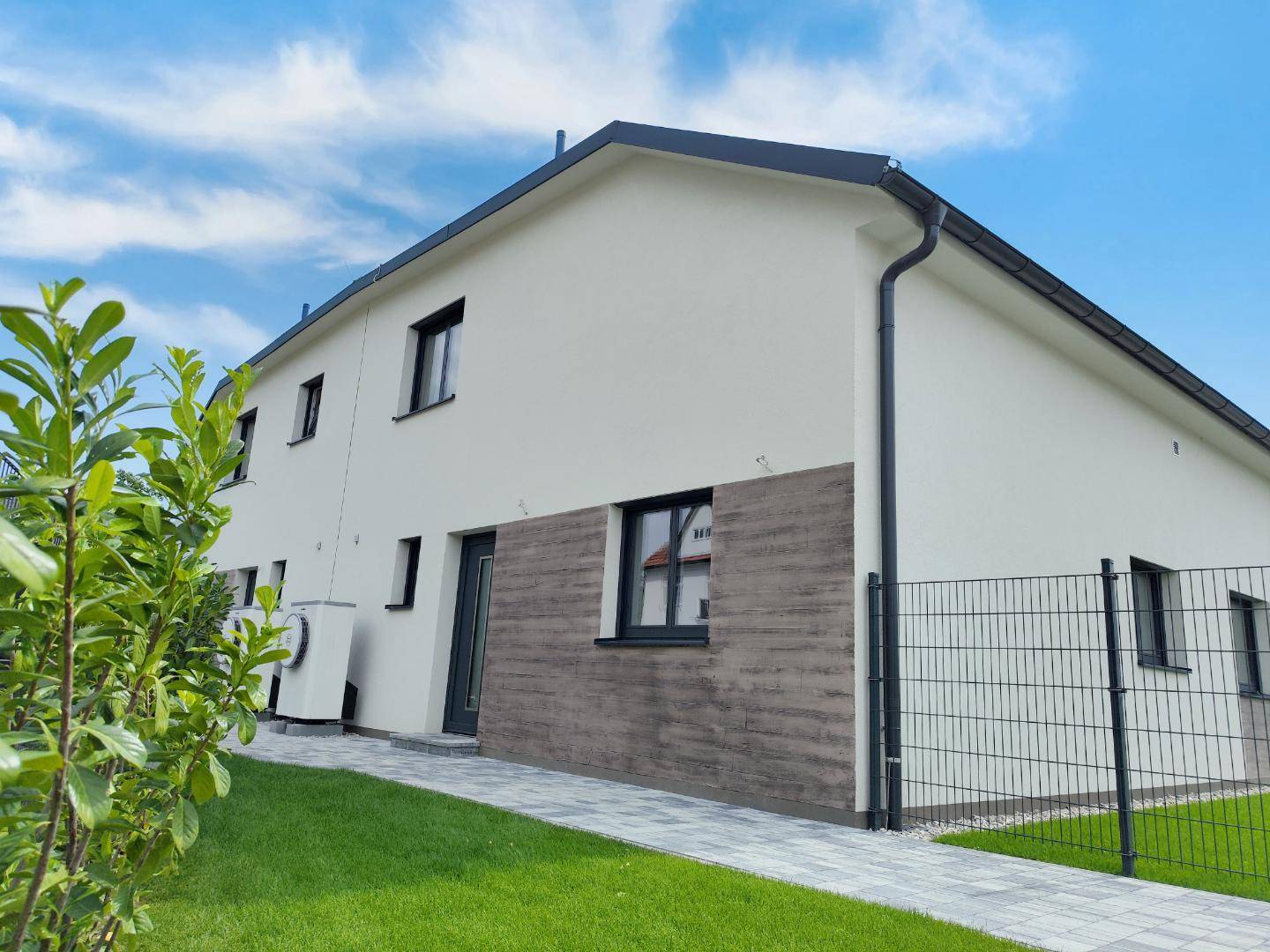 Modernes Reihenhaus mit heller Fassade, dunklem Dach und gepflegtem Rasen unter blauem Himmel.