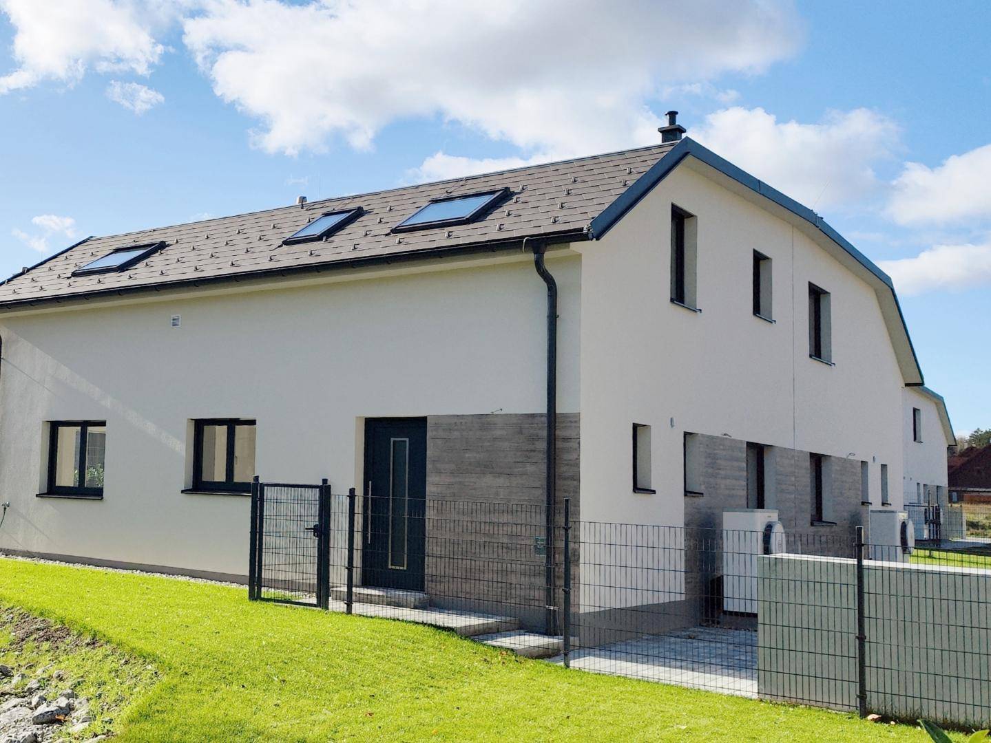 Neubau-Reihenhäuser mit heller Putzfassade, dunklen Fenstern und Dachfenstern, umgeben von gepflegtem Rasen.