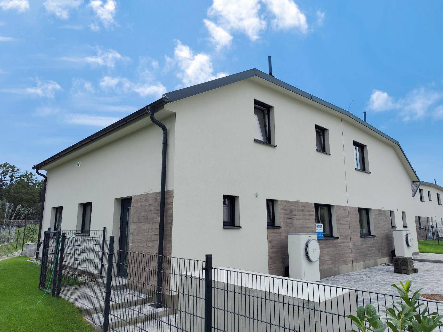 Seitenansicht eines modernen Reihenhauses mit heller Fassade, dunklem Dach und gepflastertem Zugangsbereich.
