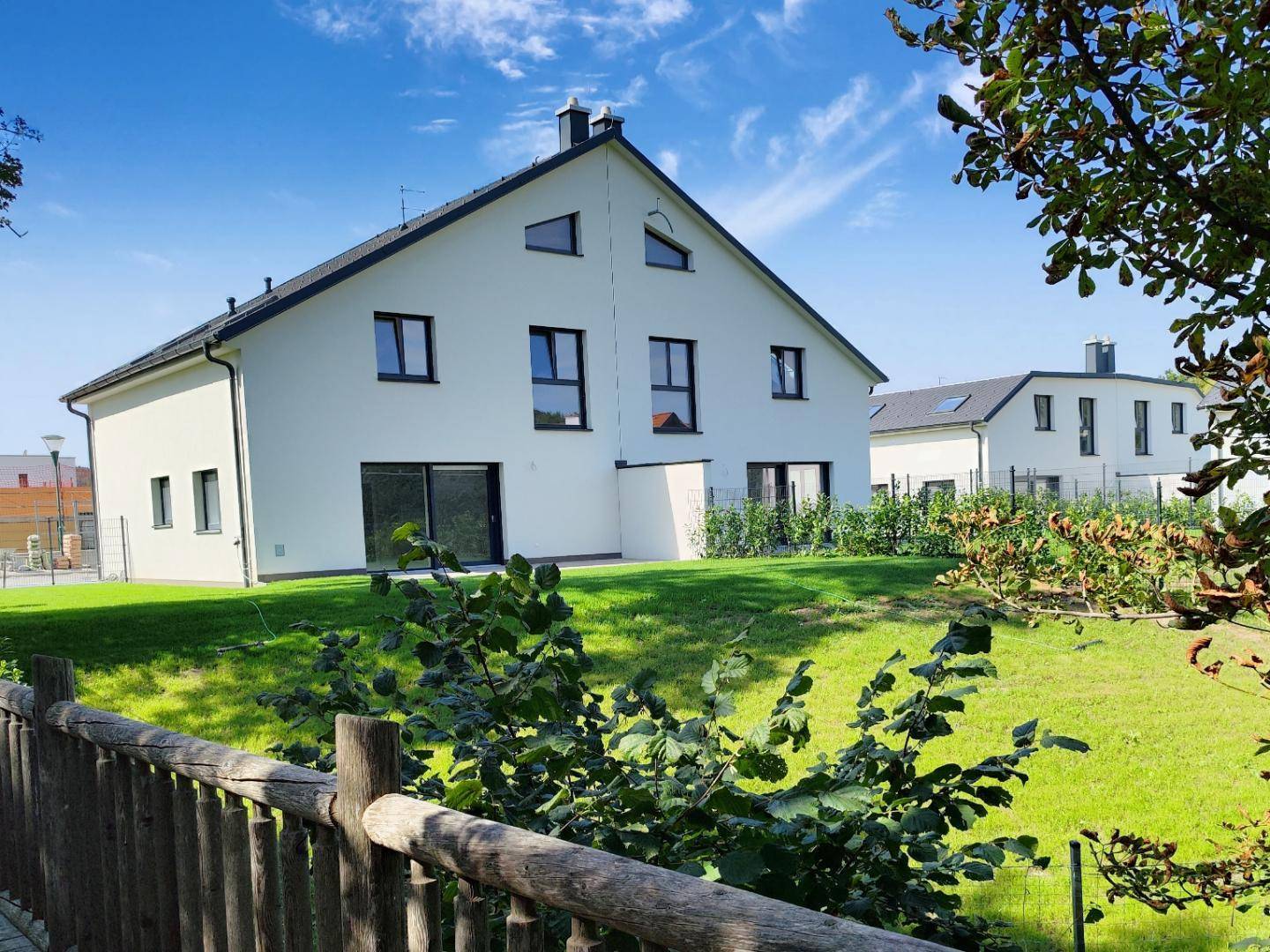 Neubau-Doppelhaushälfte mit weißer Fassade und grüner Umgebung an einem sonnigen Tag.