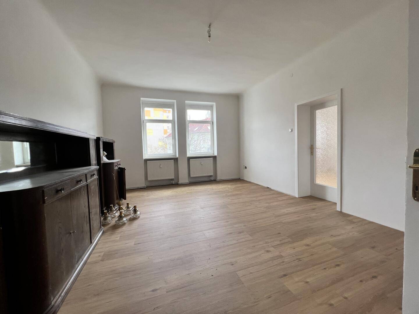 Geräumiges Wohnzimmer mit Holzboden, zwei Fenstern und antiken Möbelstücken.
