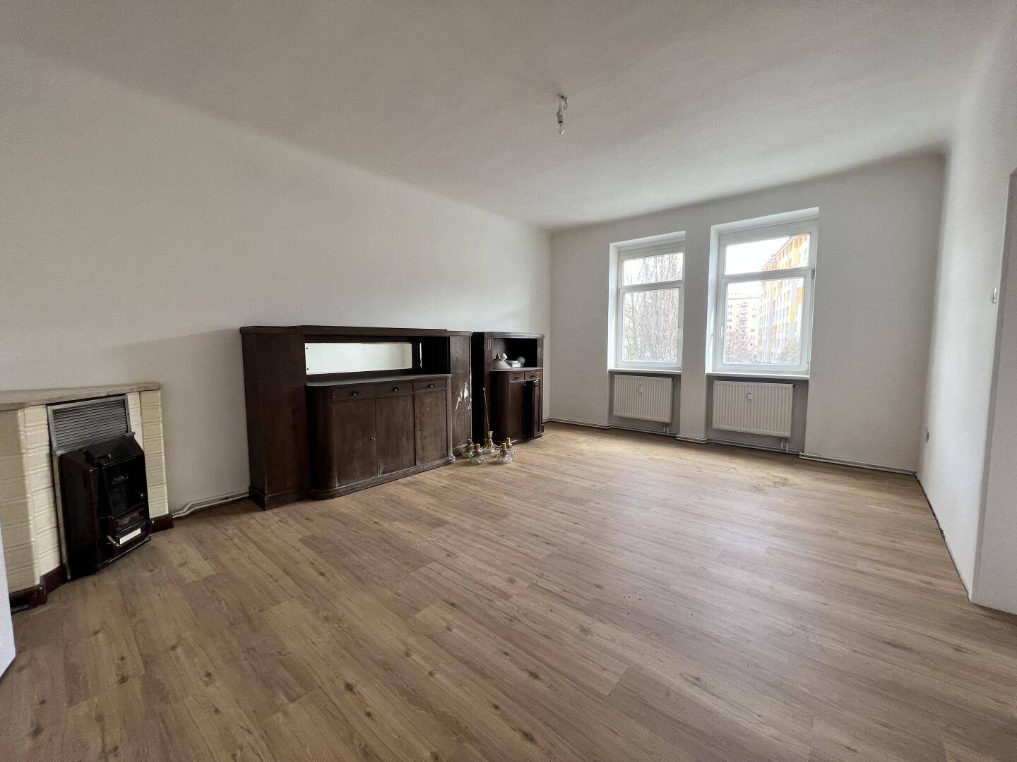 Helles Wohnzimmer mit Holzboden, zwei Fenstern und einem traditionellen Kachelofen.
