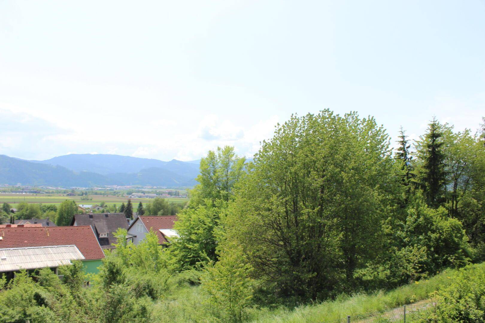 Heller Ausblick auf grüne Bäume, rote Dächer und eine weite Berglandschaft unter klarem Himmel.