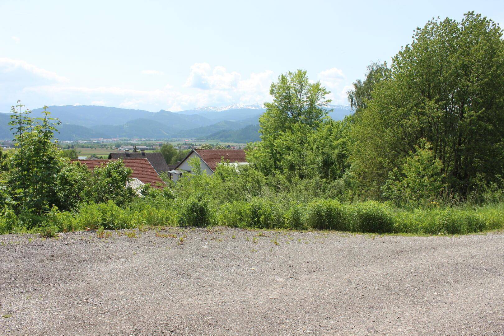 Offener Schotterbereich mit Blick auf grüne Bäume, Dächer und eine Berglandschaft.