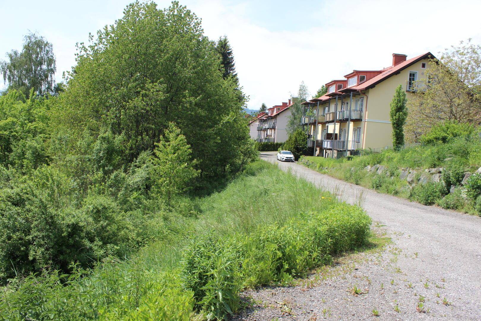 Schotterweg führt entlang eines bewachsenen Hangs zu Mehrfamilienhäusern mit Balkonen.