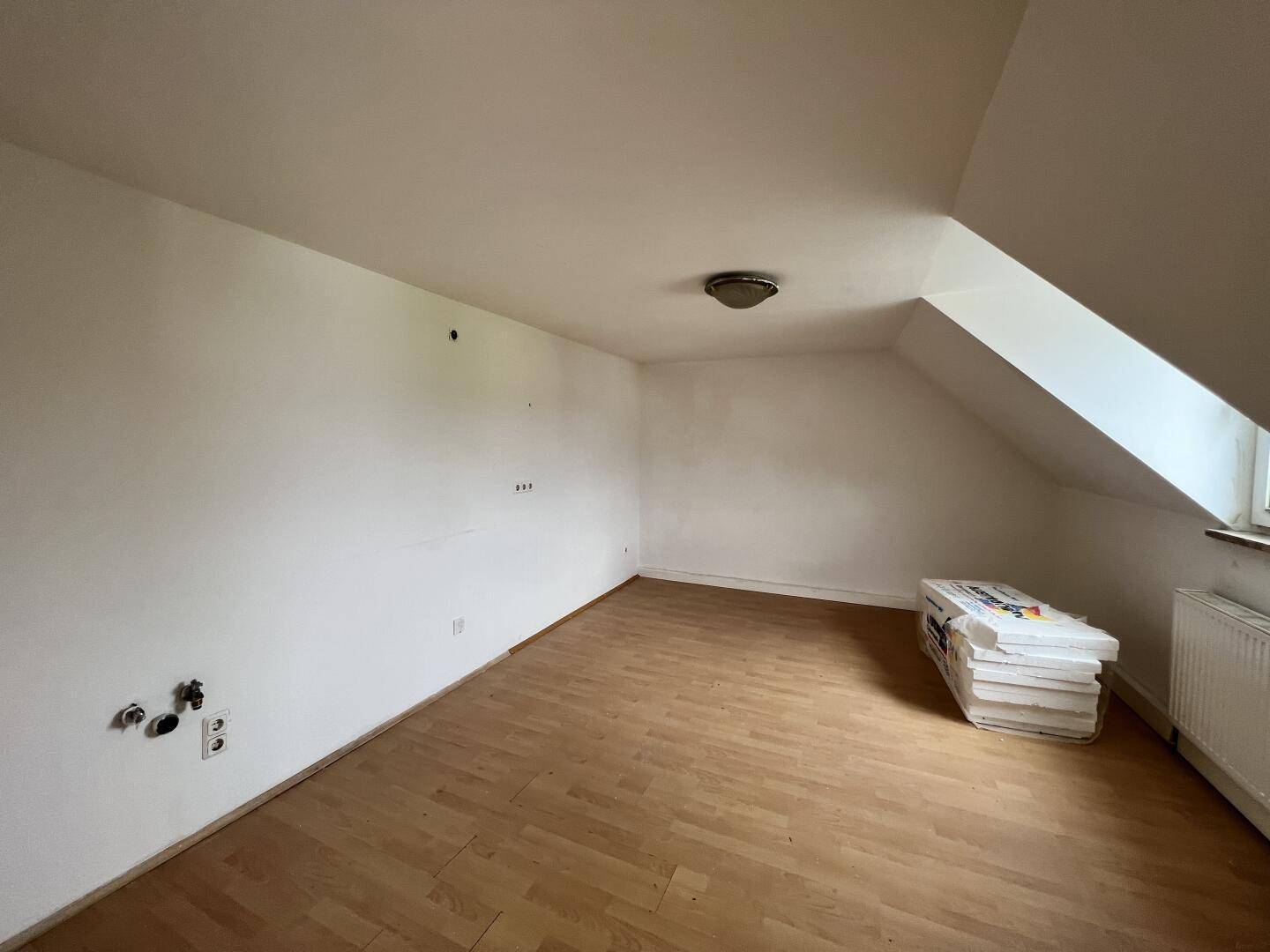 Leerer Dachbodenraum mit Laminatboden, Dachschrägen und Heizkörper unter dem Fenster.