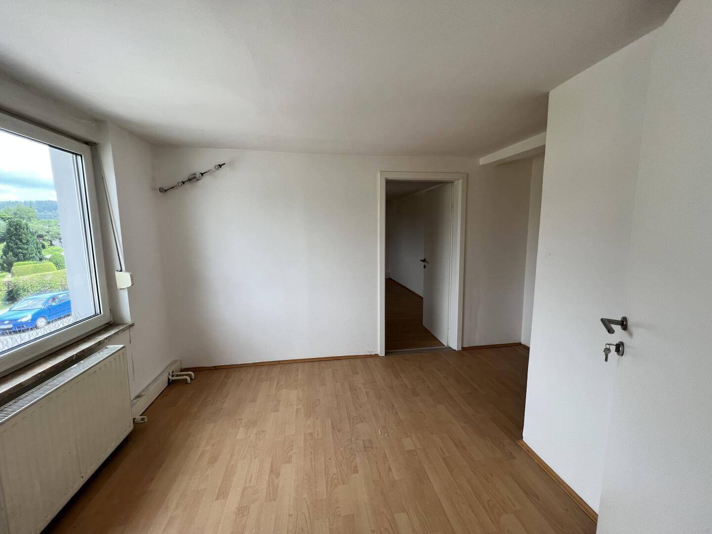 Heller, leerer Raum mit Laminatboden, großem Fenster und Zugang zu einem weiteren Zimmer.
