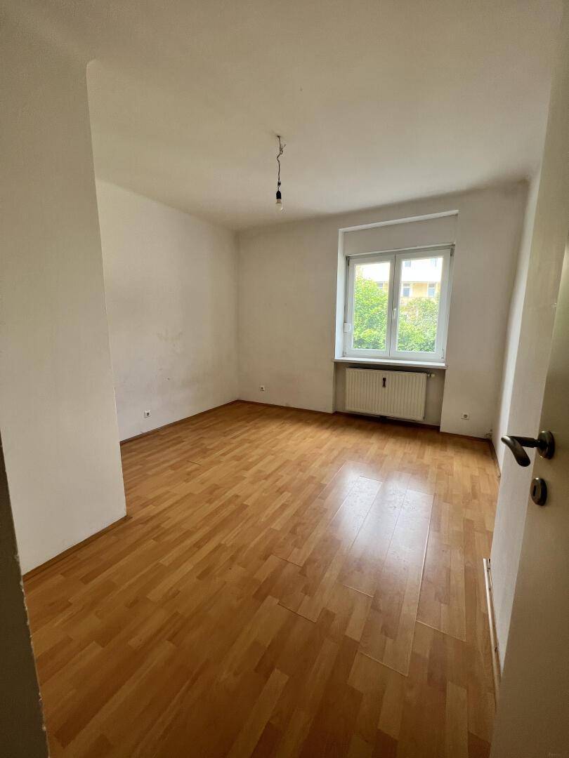 Helles, leeres Zimmer mit Parkettboden und einem Fenster, das viel Tageslicht hereinlässt.
