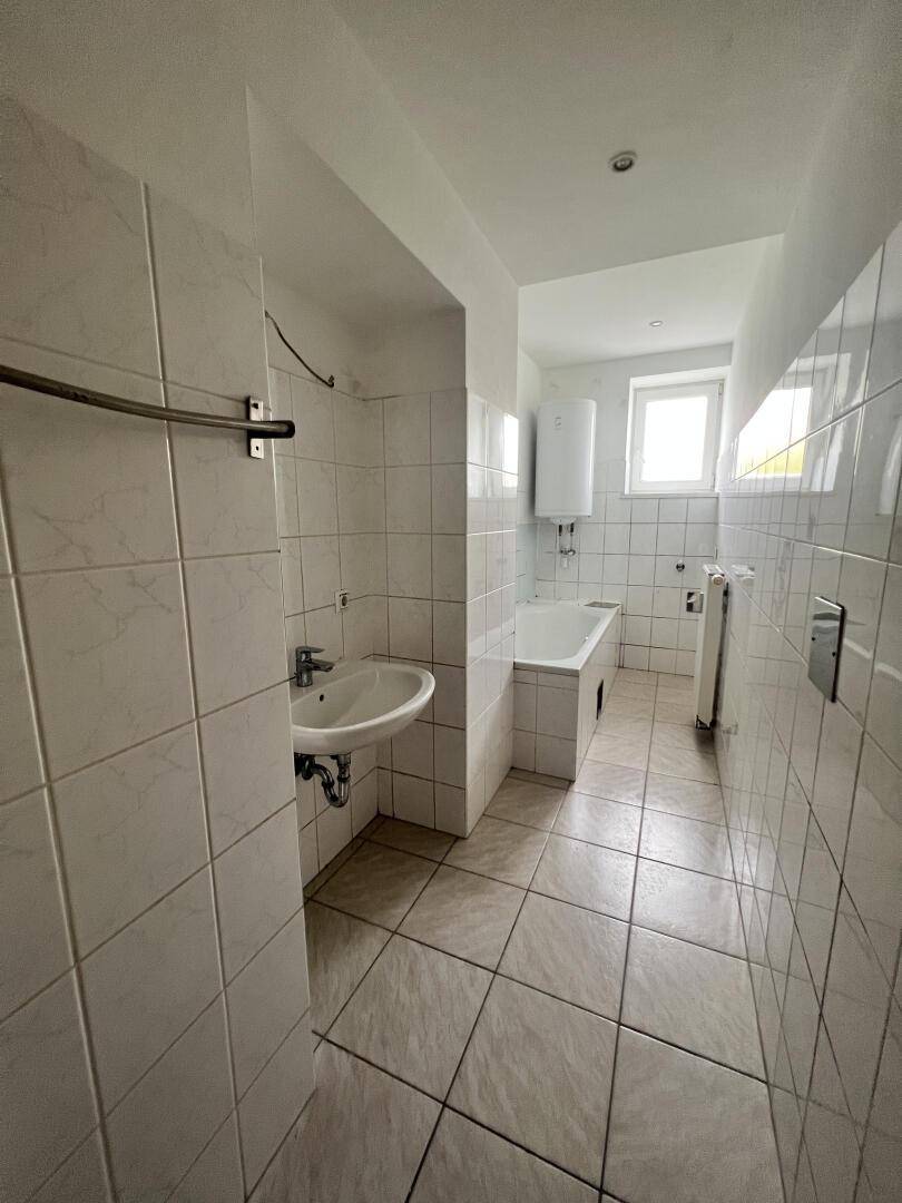 Badezimmer mit weißen Fliesen, Waschbecken, Badewanne und einem Fenster für natürliche Belüftung.