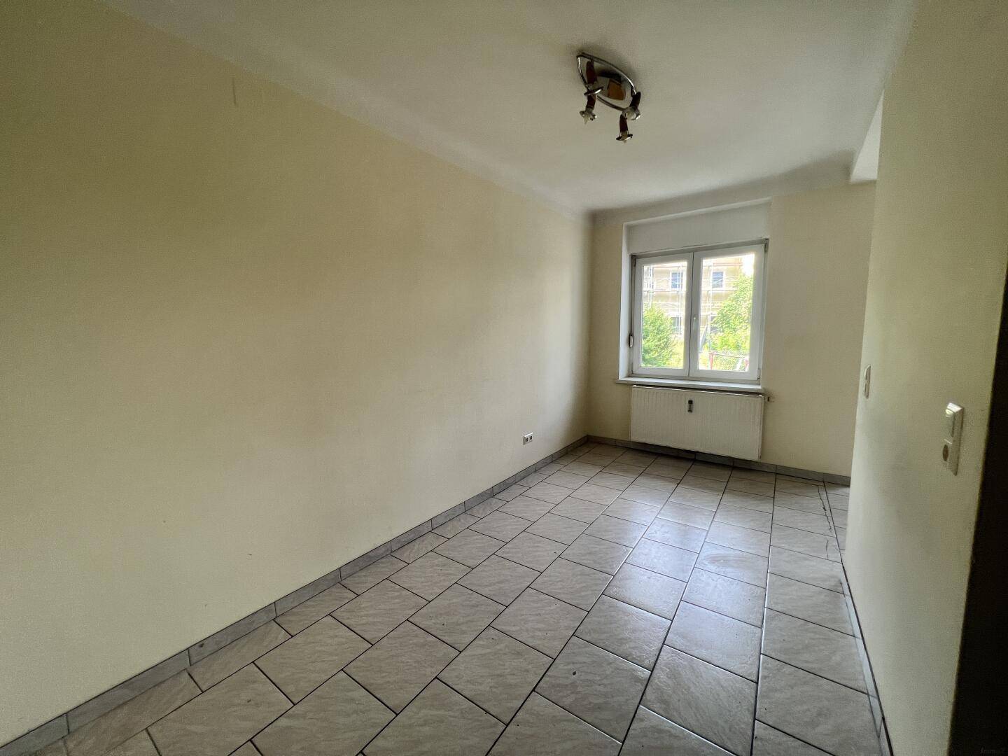Leeres Zimmer mit hellen Fliesen, einem Fenster und einem Heizkörper unter dem Fenster.