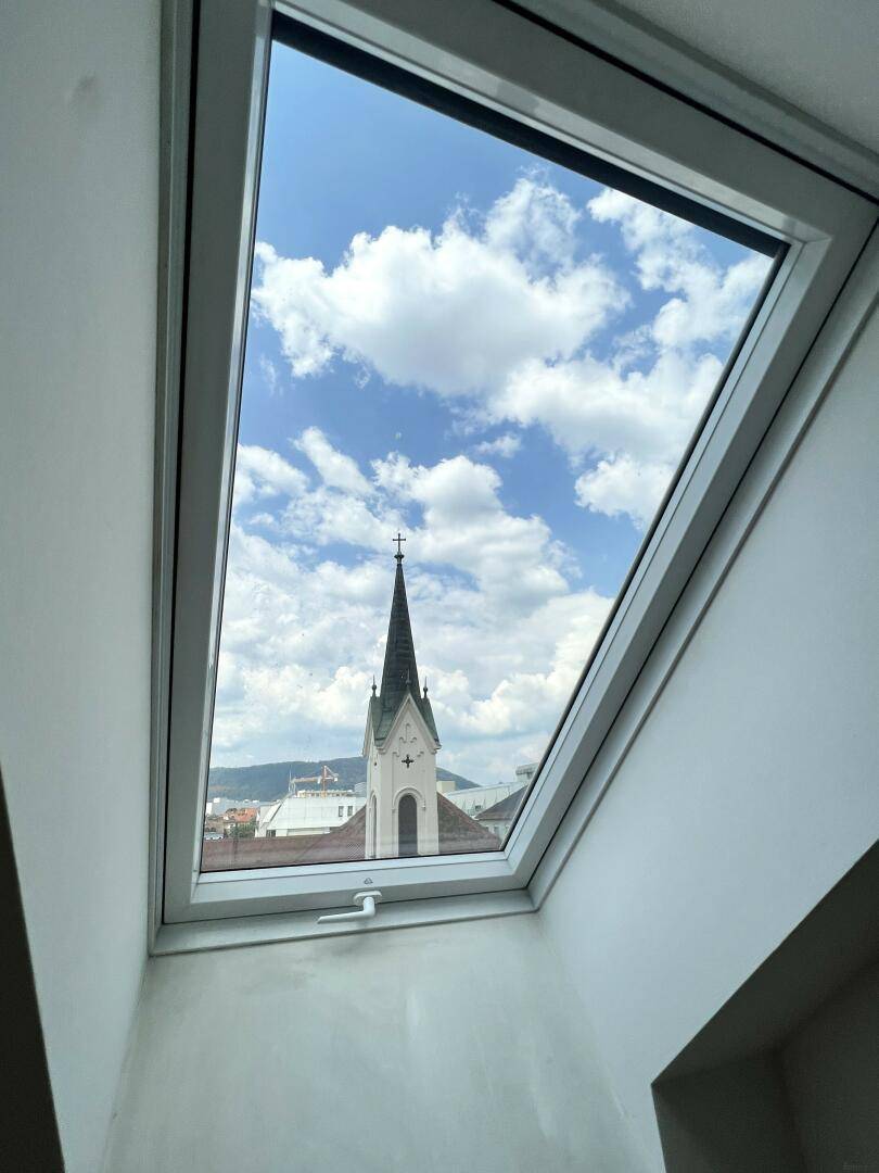 Dachfenster mit weitem Blick auf eine Kirchturmspitze und den blauen Himmel.