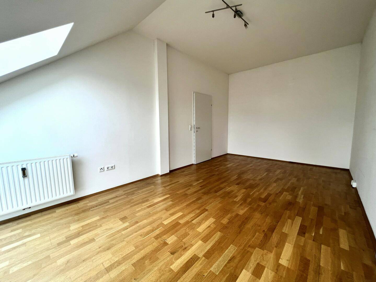 Geräumiges Zimmer im Dachgeschoss mit Parkettboden, weiß gestrichenen Wänden und Heizkörper.