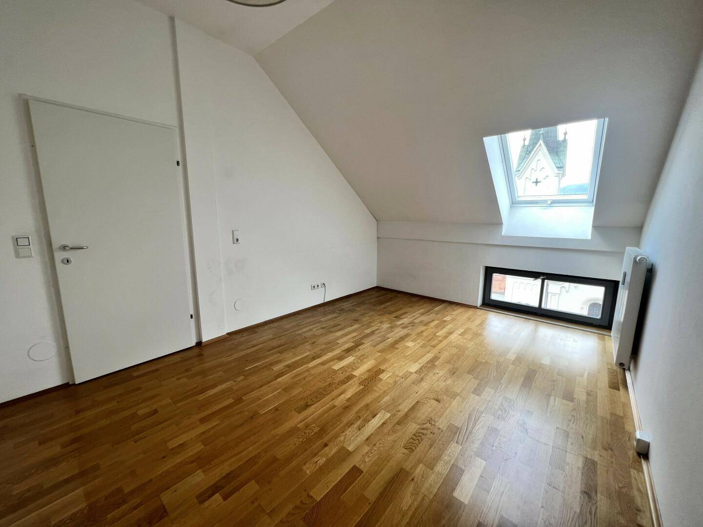Schlafzimmer im Dachgeschoss mit Parkettboden, Dachfenster und Blick auf eine Kirchturmspitze.