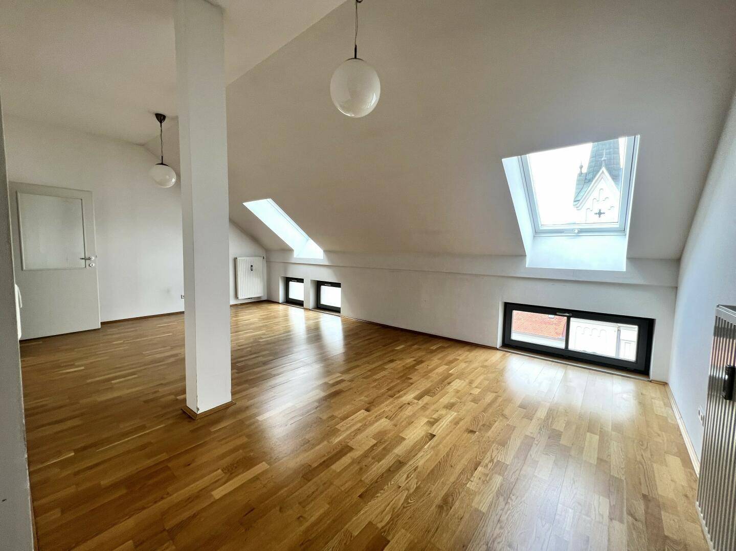 Geräumiges Dachgeschosszimmer mit Parkettboden, Dachfenstern und Blick auf eine Kirchturmspitze.