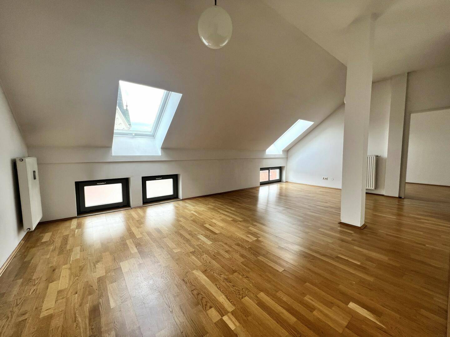 Lichtdurchfluteter Dachgeschossraum mit Holzboden, Dachfenstern und Blick auf die Umgebung.