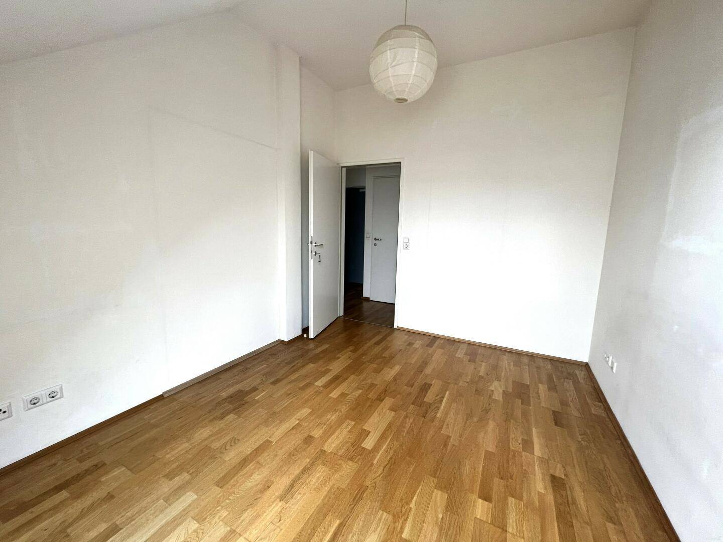 Helles Schlafzimmer mit Parkettboden, weißen Wänden und Zugang zu einem weiteren Raum.