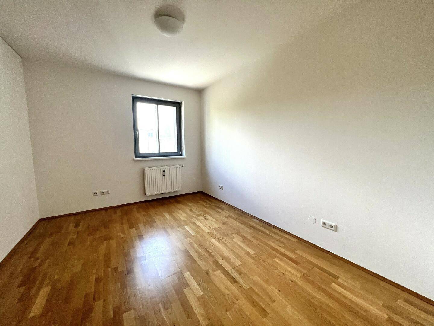 Leeres Zimmer mit hellem Parkettboden, Fenster und Heizkörper, bereit zur individuellen Gestaltung.